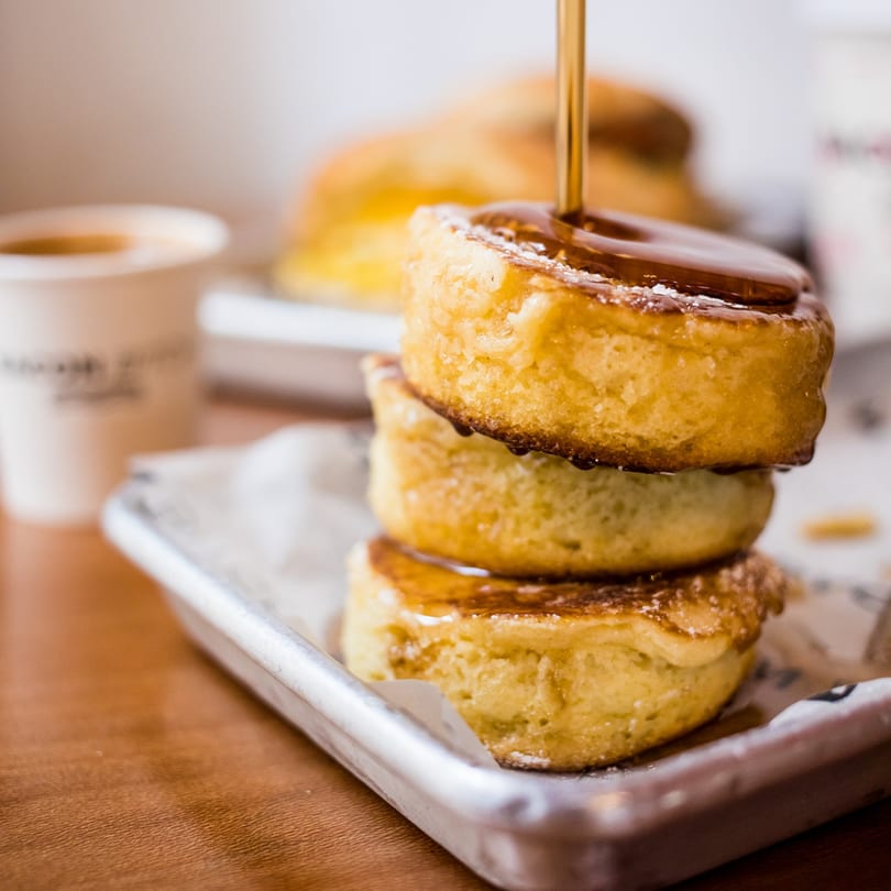 Maple syrup being poured over a stack of three fluffy pancakes dusted with powdered sugar.