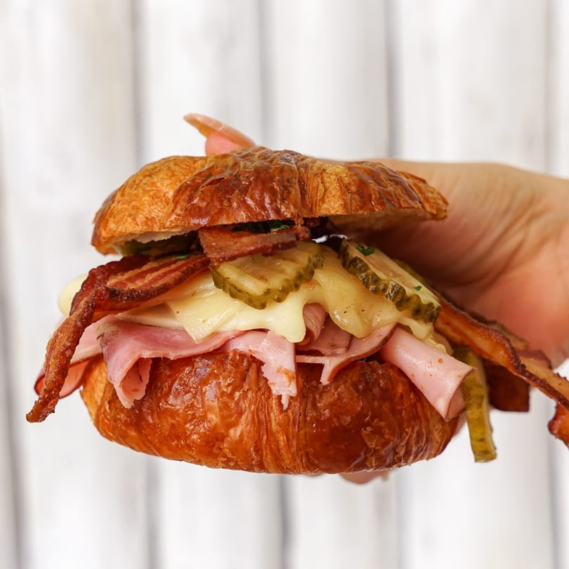 A hand holds a Bacon Bitch croissant sandwich filled with bacon, ham, melted cheese, and pickles.