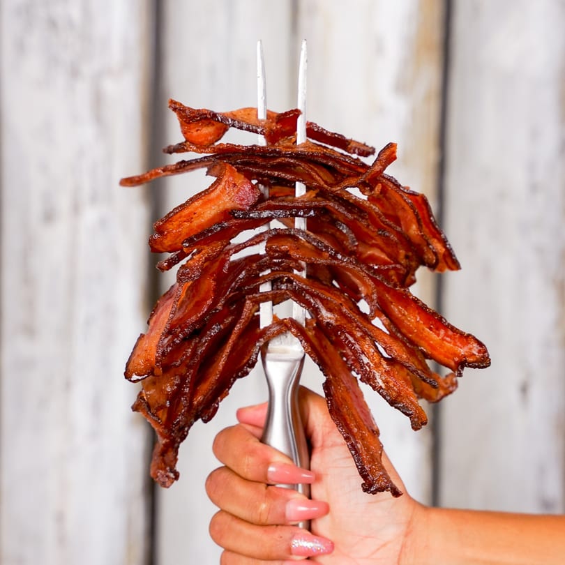 A hand holds a large fork piled high with crispy, glistening strips of Bacon Bitch bacon.