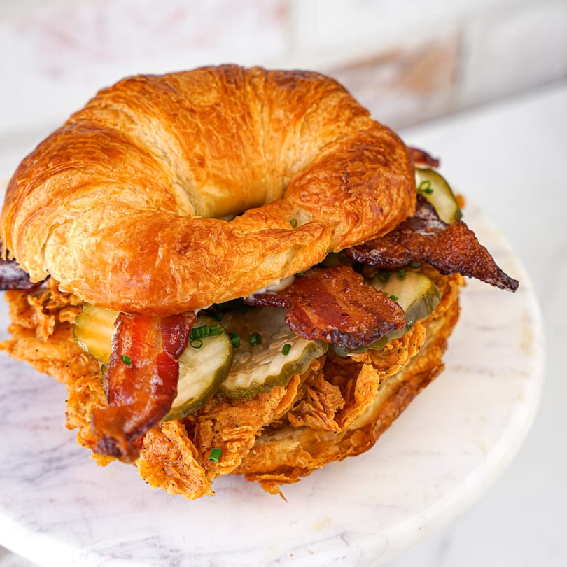 A crispy chicken sandwich with bacon, pickles, and chives on a golden croissant.