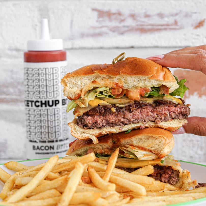 A hand holds a sliced Bacon Bitch burger with fries and a ketchup bottle in the background.