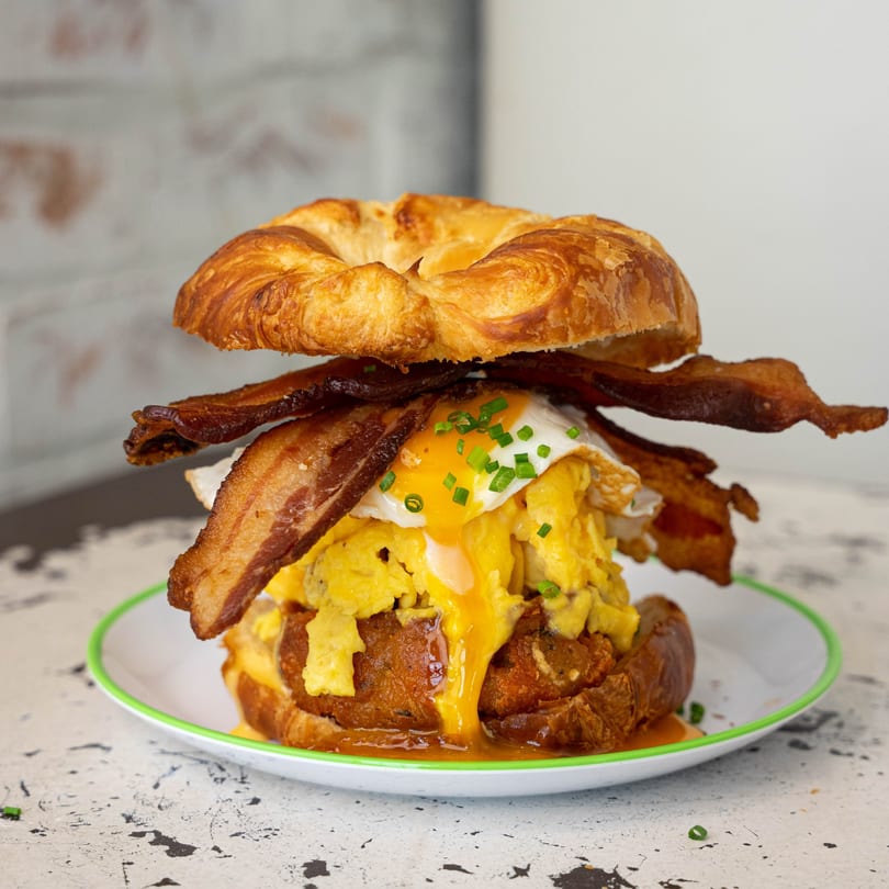 A Bacon Bitch breakfast sandwich with bacon, scrambled eggs, and a runny fried egg on a croissant.